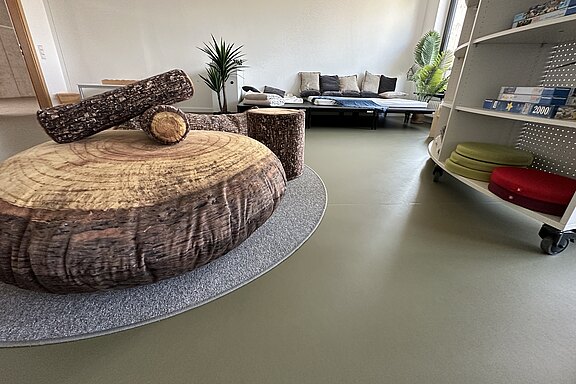 Ein moderner Raum mit einem großen runden Tisch aus Holzscheiben und einem kleinen, hölzernen Zylinder in der Mitte. Im Hintergrund steht ein gemütliches Sofa und Pflanzen sorgen für eine einladende Atmosphäre. Regale mit verschiedenen Materialien sind ebenfalls sichtbar.