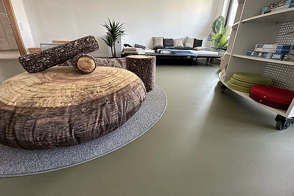 Ein moderner Raum mit einem großen runden Tisch aus Holzscheiben und einem kleinen, hölzernen Zylinder in der Mitte. Im Hintergrund steht ein gemütliches Sofa und Pflanzen sorgen für eine einladende Atmosphäre. Regale mit verschiedenen Materialien sind ebenfalls sichtbar.