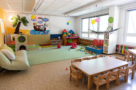Ein heller Spielraum für Kinder mit einer bunten Wandmalerei. Es gibt Spielzeuge, ein gemütliches Sofa und einen Tisch mit Stühlen. Auf dem Boden liegen bunte Spielbälle. Der Raum ist freundlich und einladend gestaltet, ideal für spielerisches Lernen und Kreativität.