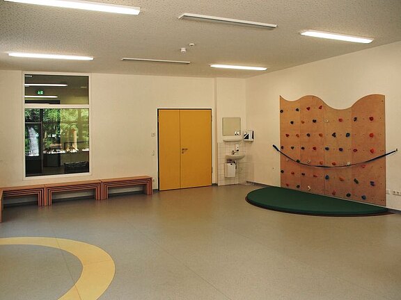 Ein leerer Raum mit hellem Boden und Wänden. Links steht eine Bank aus Holz. An der rechten Wand befindet sich eine Kletterwand mit bunten Griffen. Eine gelbe Tür ist im Mittelpunkt der Wand, und ein Waschbereich ist in der Nähe zu sehen.