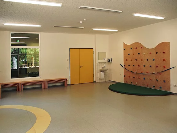Ein leerer Raum mit hellem Boden und Wänden. Links steht eine Bank aus Holz. An der rechten Wand befindet sich eine Kletterwand mit bunten Griffen. Eine gelbe Tür ist im Mittelpunkt der Wand, und ein Waschbereich ist in der Nähe zu sehen.