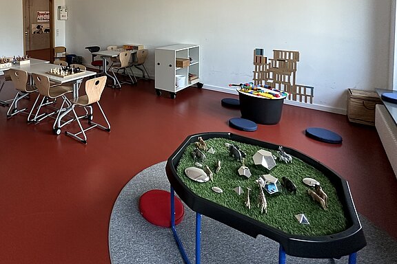 Der Raum zeigt eine Spielsituation mit einem Tisch mit Schachspielen und einem anderen Tisch für Aktivitäten. In der Mitte befindet sich ein Spieltisch mit einer grünen Fläche und verschiedenen Spielobjekten. An der Wand ist eine bunte Spielstruktur aus Holz sichtbar.