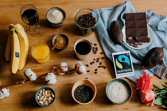 Auf einem Holztisch sind verschiedene Lebensmittel und Getränke angeordnet, darunter Bananen, Kaffeebohnen, Schokolade, Nüsse, Reis, Tee und ein Glas Orangensaft. Eine Fairtrade-Verpackung ist sichtbar. Die Anordnung vermittelt ein Gefühl von Vielfalt und Genuss.