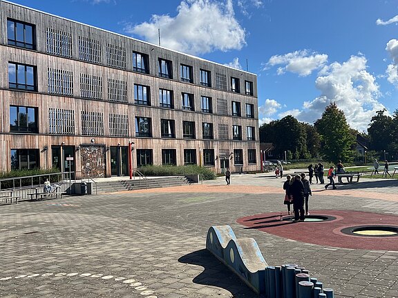 Bildbeschreibung: Eine moderne Schulgebäude mit Holzfassade steht auf einem weitläufigen Schulhof. Schüler und Schülerinnen bewegen sich auf dem Platz, der mit bunten Spielbereichen und Natur gestaltet ist. Ein klarer blauer Himmel mit wenigen Wolken ist im Hintergrund zu sehen.