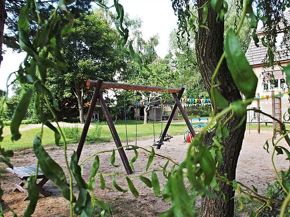 Leerer Spielplatz im Grünen: Im Vordergrund hängen Blätter, dahinter steht ein hölzernes Doppelschaukelgestell auf sandigem Boden. Im Hintergrund bunte Wimpel und weitere Spielgeräte. Sommerliche, ruhige Stimmung - vergrößerte Ansicht in Bildergalerie öffnen