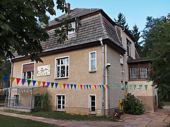 Zweistöckiges, älteres Haus, offenbar als Kindergarten genutzt. An der Fassade ein kindlich gestaltetes Schild; quer über den Hof hängen bunte Wimpelketten. Sommerliche Außenansicht mit etwas Grün und Bäumen - vergrößerte Ansicht in Bildergalerie
öffnen