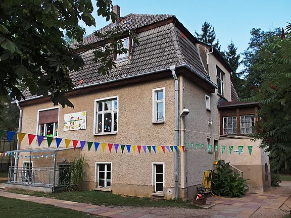 Zweistöckiges, älteres Haus, offenbar als Kindergarten genutzt. An der Fassade ein kindlich gestaltetes Schild; quer über den Hof hängen bunte Wimpelketten. Sommerliche Außenansicht mit etwas Grün und Bäumen - vergrößerte Ansicht in Bildergalerie
öffnen
