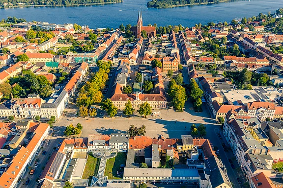 Luftaufnahme einer Stadt mit einem Marktplatz in der Mitte. Umgeben von bunten Gebäuden und Bäumen, führt eine Straße zu einem Fluss im Hintergrund. Ein markantes Kirchenschiff ragt in die Höhe und prägt das Stadtbild.