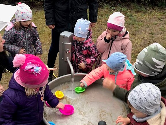 Eine Gruppe von Kindern steht um ein Wasserbecken im Freien. Eine erwachsene Person, die sie betreut, lächelt sie an. Die Kinder spielen mit bunten Bechern im Wasser und tragen warme Kleidung. Der Hintergrund zeigt eine eingezäunte Grünfläche.