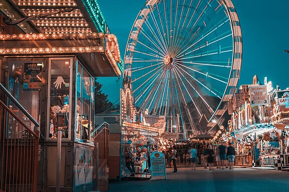 Abendliche Kirmesszene: Ein hell beleuchtetes Riesenrad dominiert den Hintergrund, rechts und links stehen bunte Buden mit Lichtern. Menschen schlendern über den Jahrmarkt, verschwommen in Bewegung. Die Atmosphäre wirkt lebhaft und stimmungsvoll.