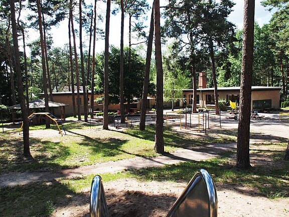 Ein Spielplatz in einem Waldgebiet mit Holzhütten und Spielgeräten. Die Umgebung ist grün und sonnig, umgeben von hohen Bäumen. Im Hintergrund sind Sitzgelegenheiten und Spielmöglichkeiten sichtbar, die zu einem freundlichen und einladenden Spieleplatz beitragen.
