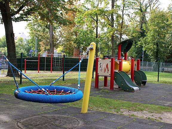 Spielplatz im Freien: Im Vordergrund eine blaue Nestschaukel, dahinter ein kleines Kletter- und Rutschspielgerät mit Röhre und zwei Rutschen. Der Bereich ist umzäunt und von Bäumen umgeben.