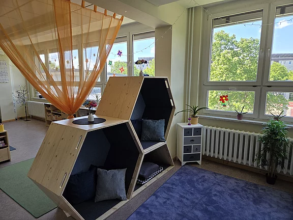 Heller Raum mit zwei übereinander angeordneten, sechseckigen Holznischen mit dunkler Polsterung und Kissen – eine gemütliche Lese- oder Ruheecke. Daneben große Fenster, etwas Grünpflanzen und ein kleines Schränkchen. Insgesamt wirkt der Bereich freundlich und einladend.
