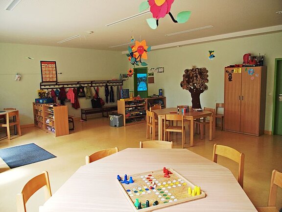 Ein heller, einladender Kindergartenraum mit einem großen Tisch in der Mitte, auf dem ein Brettspiel liegt. Um den Raum sind Tische und Stühle angeordnet, mit Regalen für Spielsachen und Kleidung. Bunte Dekorationen und Spielzeuge schaffen eine fröhliche Atmosphäre.