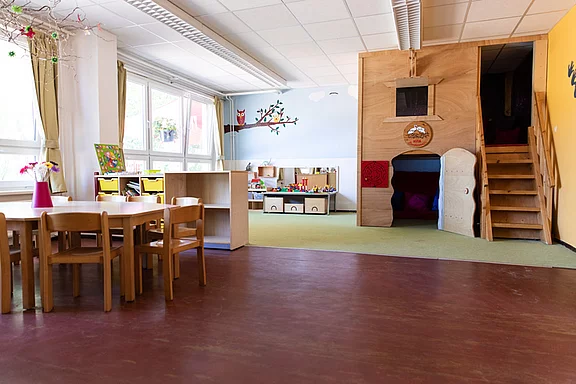 Der Raum ist ein helles Klassenzimmer mit einem großen Tisch aus Holz und Stühlen. An der Wand befindet sich ein Baum mit einer Eule. In der Ecke steht ein Spielbereich mit Kisten und Spielzeug. Rechts gibt es einen aufgebauten Spielplatz mit einer kleinen Treppe.
