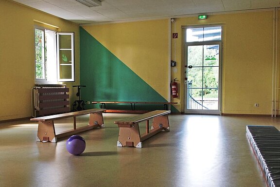 Ein heller Raum mit gelben Wänden und einem grünen Akzent. Auf dem Boden stehen zwei Turnbänke sowie ein lila Ball. Ein offenes Fenster lässt Tageslicht herein, während die Tür zum Außenbereich sichtbar ist.