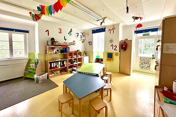 Heller Kindergartenraum: kleiner Tisch mit Hockern zentral, Zahlen 1–12 an den Wänden, Regale mit Spielen und Büchern, bunte Deckendeko. Viel Tageslicht durch Fenster und Tür; Raum fürs frühe Lernen, Basteln und Spielen.