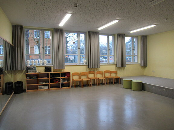 Ein heller Raum mit großen Fenstern und Vorhängen. An einer Wand stehen Regale mit Geräten, daneben sind zwei schwarze Lautsprecher. Im Raum befinden sich mehrere Holzstühle und zwei grüne Sitzwürfel. Ein Spiegel reflektiert die gegenüberliegende Wand.