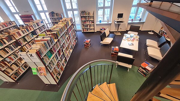 Blick von einer Wendeltreppe in einen hellen Bibliotheksraum: hohe Fenster, lange Reihen gut gefüllter Bücherregale, daneben eine kleine Leseecke mit Sofas, Tisch und Computer. Bücherstapel stehen auf dem Boden.