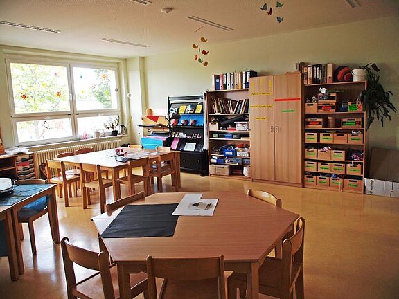 Ein heller Klassenraum mit mehreren runden und rechteckigen Tischen, Stühlen und Regalen. An den Wänden hängen bunte Dekorationen. Im Hintergrund sind Fenster, die Tageslicht hereinlassen, sowie Bücherregale mit verschiedenen Materialien und Bastelsachen.