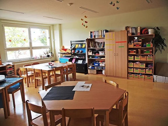 Ein heller Klassenraum mit mehreren runden und rechteckigen Tischen, Stühlen und Regalen. An den Wänden hängen bunte Dekorationen. Im Hintergrund sind Fenster, die Tageslicht hereinlassen, sowie Bücherregale mit verschiedenen Materialien und Bastelsachen.