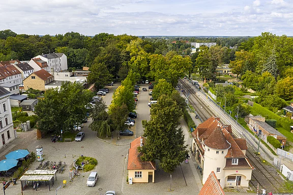 Luftaufnahme des Bahnhofes Neuruppin Rheinsberger Tor, rechts verlaufen Gleise neben einem historischen Stationsgebäude mit roten Ziegeldächern, links ein Parkplatz mit zahlreichen Autos und ein Platz mit Fahrrädern und wenigen Passanten, umgeben von der Stadtmauer, dichten Bäumen und historisch gestalteten Wohnhäusern.