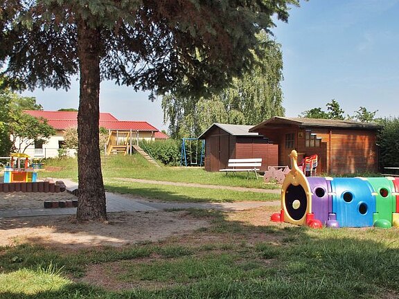Ein Spielplatz im Freien mit einem bunten Spielgerät in Form eines Zuges. Im Hintergrund stehen ein Holzhaus und eine Schaukel. Umgeben von Rasen und Bäumen, strahlt die Szene eine einladende, kinderfreundliche Atmosphäre aus.