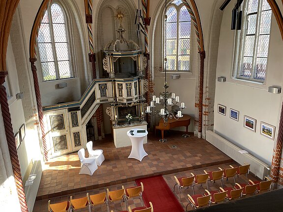 Man sieht den Innenraum einer Kapelle mit hohen Fesntern, durch die die Sonne scheint. Man schaut von der Empore hinunter und erkennt einen Stehtisch, auf dem eine Mappe für die Eheschließung liegt. Links davon befinden sich zwei Stühle, auf denen das Brautpaar Platz nehmen kann. Davor sind Stuhlreihen für die Gäste aufgebaut.