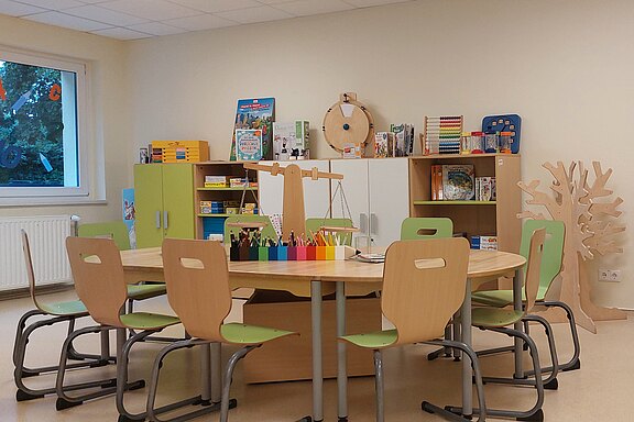 Heller Klassenraum mit großem Gruppentisch und kleinen Stühlen. Auf dem Tisch bunte Stifte und Bastelmaterial. Regale mit Büchern und Spielen, Globus auf Tisch und hängender Erdball. Freundliche Lern- und Bastelumgebung für Kinder.