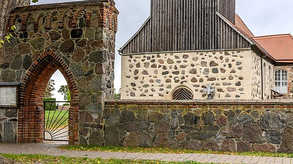 Ein historisches Steintor aus Backstein und großen Granitsteinen führt zu einem Gebäudekomplex mit einer Mischung aus hellen und dunklen Mauern sowie einem hohen Holzturm. Der Eingang ist von einer gepflasterten Fläche und einer grünen Wiese umgeben.