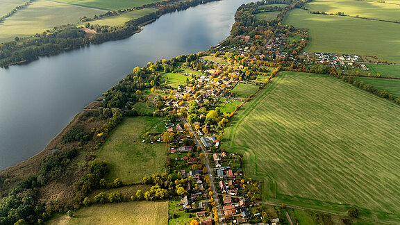 Luftaufnahme eines ländlichen Gebiets mit einem Fluss, der durch die Landschaft fließt. Auf der einen Seite sind Wiesen und Felder, auf der anderen Seite eine kleine Ansiedlung mit Wohnhäusern und Bäumen. Die Natur zeigt herbstliche Farben.