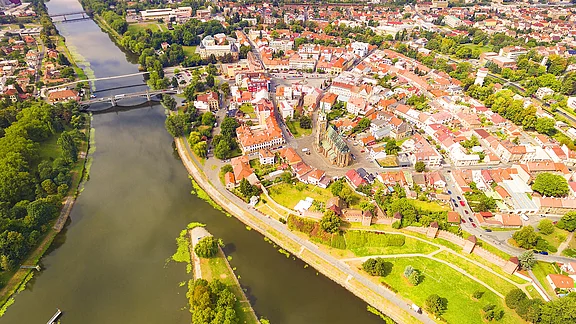Luftaufnahme einer Stadt, die sich entlang eines Flusses erstreckt. Zu sehen sind eine Vielzahl von Gebäuden mit roten Dächern, grüne Parks und Straßen. Im Zentrum der Stadt steht eine Kirche mit einem hohen Turm. Der Fluss fließt ruhig daran vorbei.