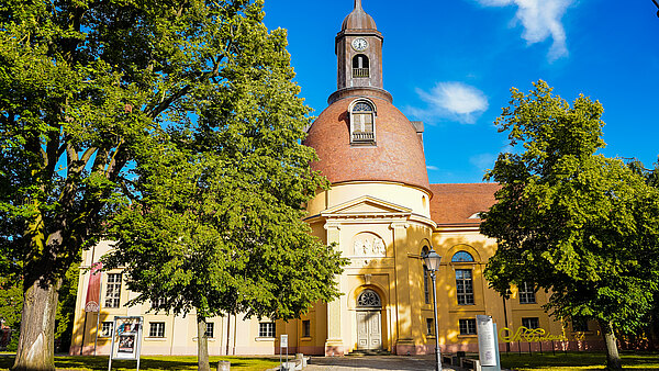 Eine gelbe Kirche mit einer großen, runden Kuppel und einem Glockenturm steht inmitten von grünen Bäumen. Der Himmel ist blau mit einigen Wolken. Die Fassade der Kirche zeigt architektonische Details, einschließlich einer Haupttür und Fenstern.