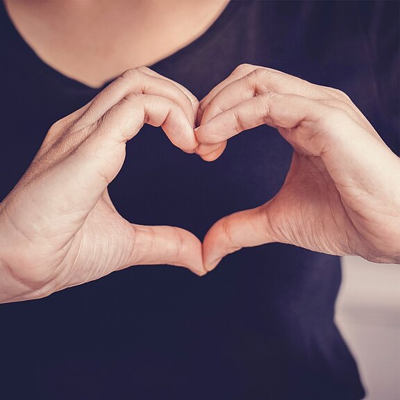 Zwei Hände formen ein Herzsymbol, das die Darstellung von Liebe und Zuneigung vermittelt. Der Hintergrund ist unscharf, sodass der Fokus auf den Händen liegt.
