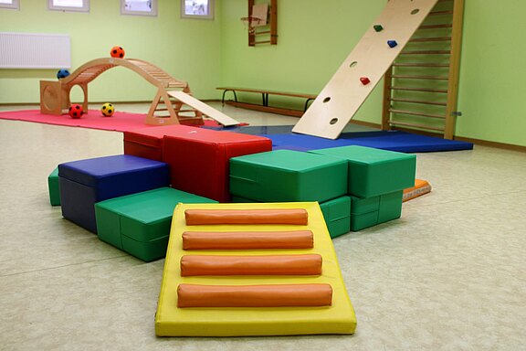 Heller Innenraum für Kinderturnen: bunte Schaumstoffblöcke und Matten, eine weiche Stufenrampe, schiefe Kletterebene mit Griffsteinen, Balancierbogen. Gepolsterte Geräte bilden einen Parcours zum Klettern, Krabbeln und Balancieren.
