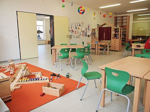 Ein helles Klassenzimmer mit Tischen und grünen Stühlen. Auf dem Boden liegt ein orangefarbenes Spielteppich mit Spielzeugautos und Bauklötzen. An den Wänden hängen Zeichnungen von Kindern. Die Atmosphäre ist einladend und fördert kreatives Spiel und Lernen.