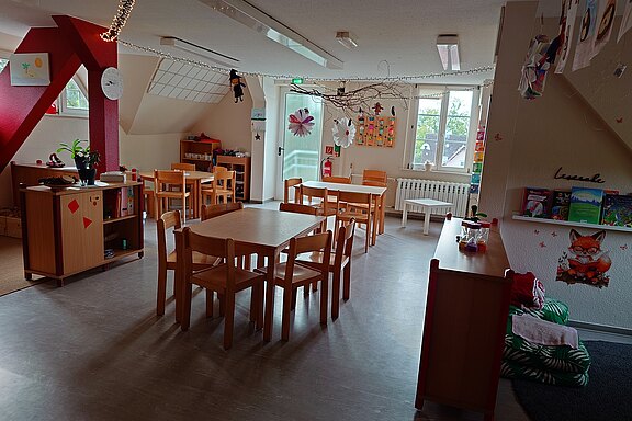Heller Kindergartenraum: mehrere kleine Holztische mit Stühlen, Regale mit Büchern und Materialien, viele Kinderzeichnungen an Wänden und von der Decke. Große Fenster bringen Tageslicht. Rechts eine gemütliche Lese- und Kuschelecke. Aufgeräumte, freundliche Lern- und Spielumgebung.