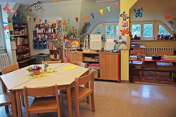 Ein helles Klassenzimmer mit einem runden Tisch und Stühlen in der Mitte. Bunte Girlanden und Schmetterlinge schmücken die Wände. An einer Wand stehen Regale mit Büchern und Materialien, während auf einer anderen Seite Maleasel und Bastelmaterialien bereitliegen.