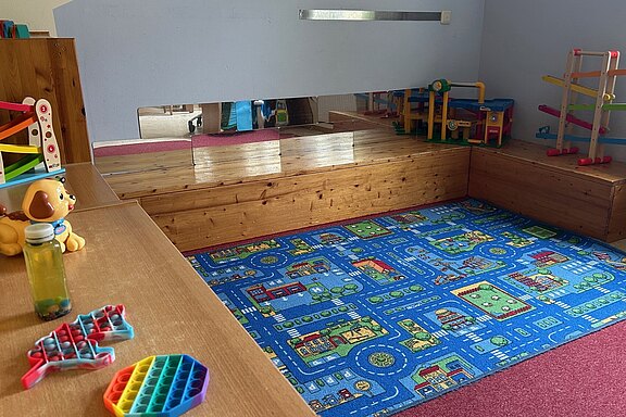 Im Bild ist ein Spielzimmer zu sehen. Der Boden ist mit einem bunten Teppich in Straßen- und Hausdesign gestaltet. Auf einem Tisch liegen einige Spielzeuge, darunter ein Hund und zwei Spielgeräte. An einer Wand befinden sich Regale mit weiteren Spielzeugen.