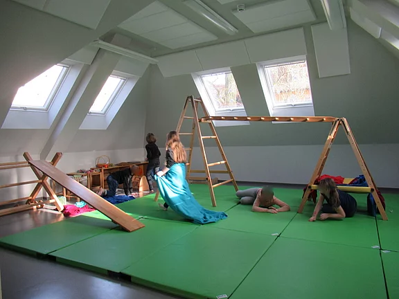 In einem hellen Raum mit schrägen Fenstern spielen Kinder auf grünen Matten. Eine Leiter und eine Schaukel aus Holz stehen im Hintergrund. Ein Mädchen in einem blauen Kleid bewegt sich, während andere Kinder liegen oder sich auf den Matten aufhalten.