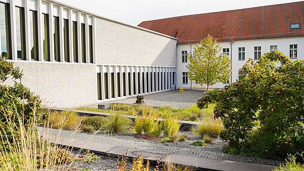 Im Fokus der Szene steht ein moderner Gebäudekomplex mit großen Fenstern, umgeben von einem angelegten Garten mit bunten Pflanzen und schmalen Wegen. Ein grüner Baum und eine kleine Statuette sind ebenfalls sichtbar. Der Garten vermittelt eine ruhige und einladende Atmosphäre.
