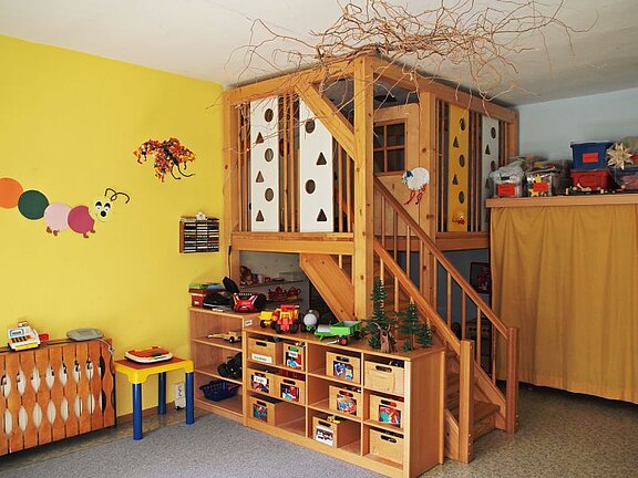 Ein helles Spielzimmer mit gelben Wänden. In der Ecke steht eine holzfarbene Spielburg mit einer Treppe. Vor der Spielburg befindet sich ein Regal mit vielen bunten Spielzeugen. Auf den Wänden sind kindgerechte Bilder zu sehen, einschließlich einer bunten Raupe.