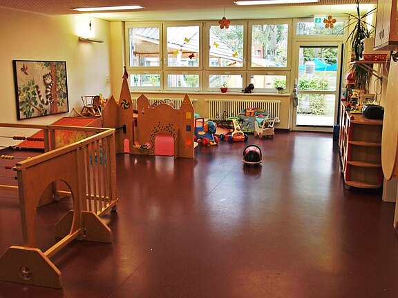 Ein heller Spielraum mit Holzspielgeräten, darunter eine kleine Kletterstruktur und Spielzeuge wie Fahrzeuge und Puppen. Große Fenster bieten natürlichen Lichtblick auf den Außenbereich. Der Boden ist glatt und rot, die Wände sind in warmen Farben gehalten.