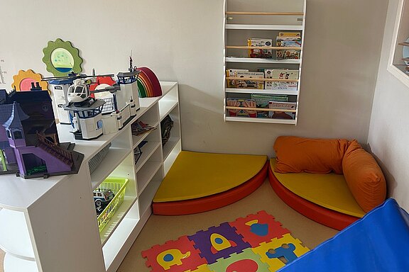 Ein heller Spielraum mit einer Spielecke. Auf einer weißen Regalwand stehen Spielzeug und Bücher. In der Ecke befinden sich zwei gepolsterte Sitzflächen, eine gelb und eine rot. Der Boden ist mit einem bunten Kinderteppich mit Buchstabenmuster ausgestattet.