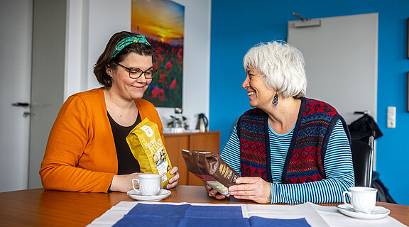 Zwei Frauen sitzen an einem Tisch in einem hellen Raum. Sie lächeln sich an und halten jeweils eine Tüte mit Lebensmitteln. Auf dem Tisch stehen zwei Kaffeetassen und eine Tischdecke mit blauem und weißem Muster.