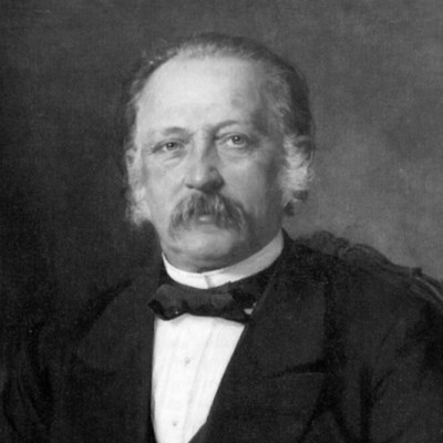 Ein schwarz-weißes Porträt eines Mannes mit einem Schnurrbart und einem Anzug. Er hat einen ernsten Gesichtsausdruck und schaut direkt in die Kamera. Der Hintergrund ist verschwommen und trägt zur fokussierten Darstellung des Mannes bei.